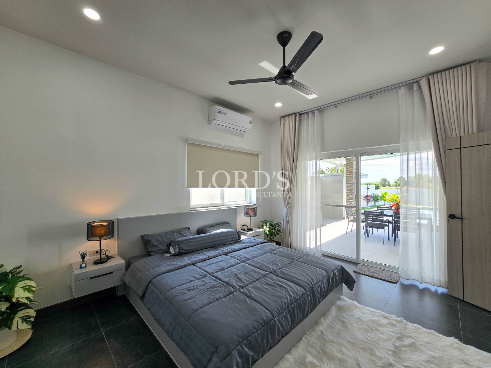 Modern bedroom with gray bedding, ceiling fan, wall-mounted air conditioner, and sliding glass doors opening to a patio.