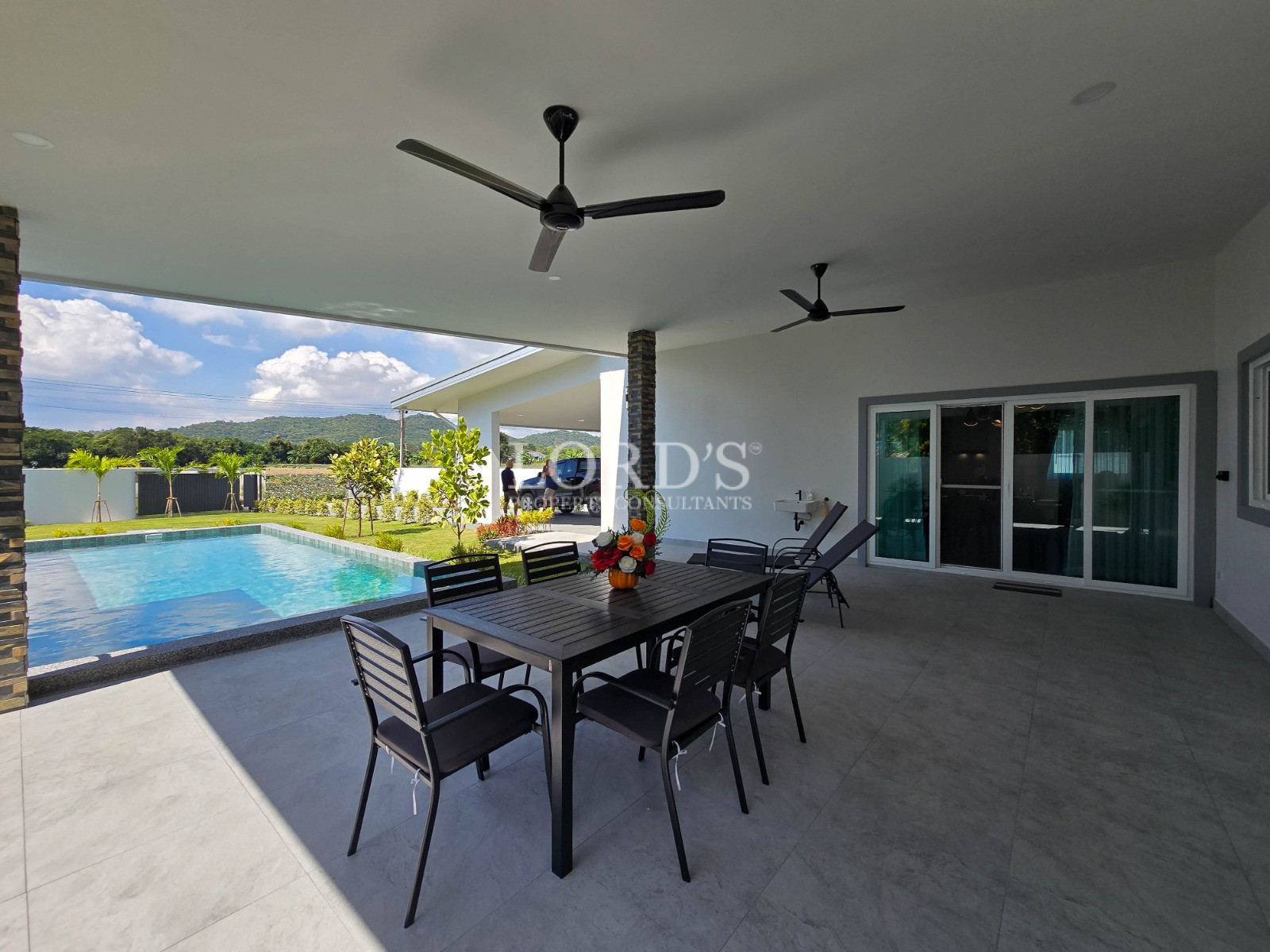 Covered outdoor patio with dining table and chairs overlooking a swimming pool and landscaped garden.