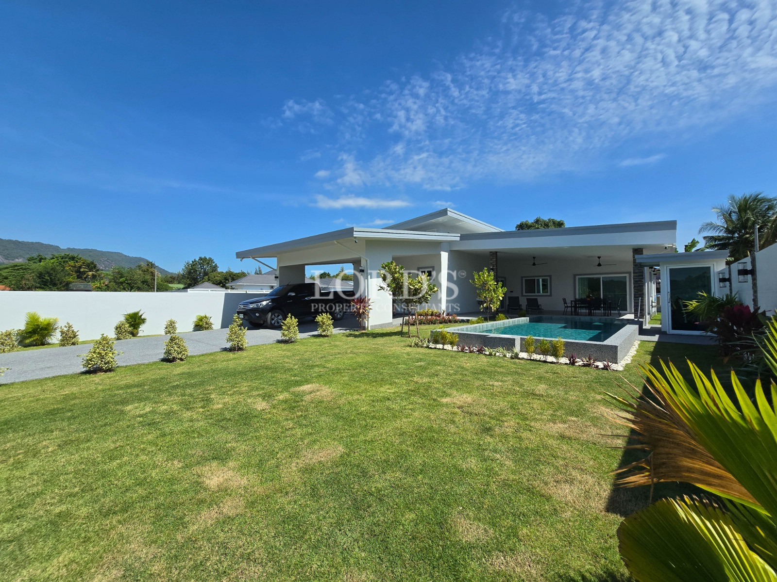 Modern single-story home with carport, landscaped lawn, and private swimming pool under a bright blue sky.