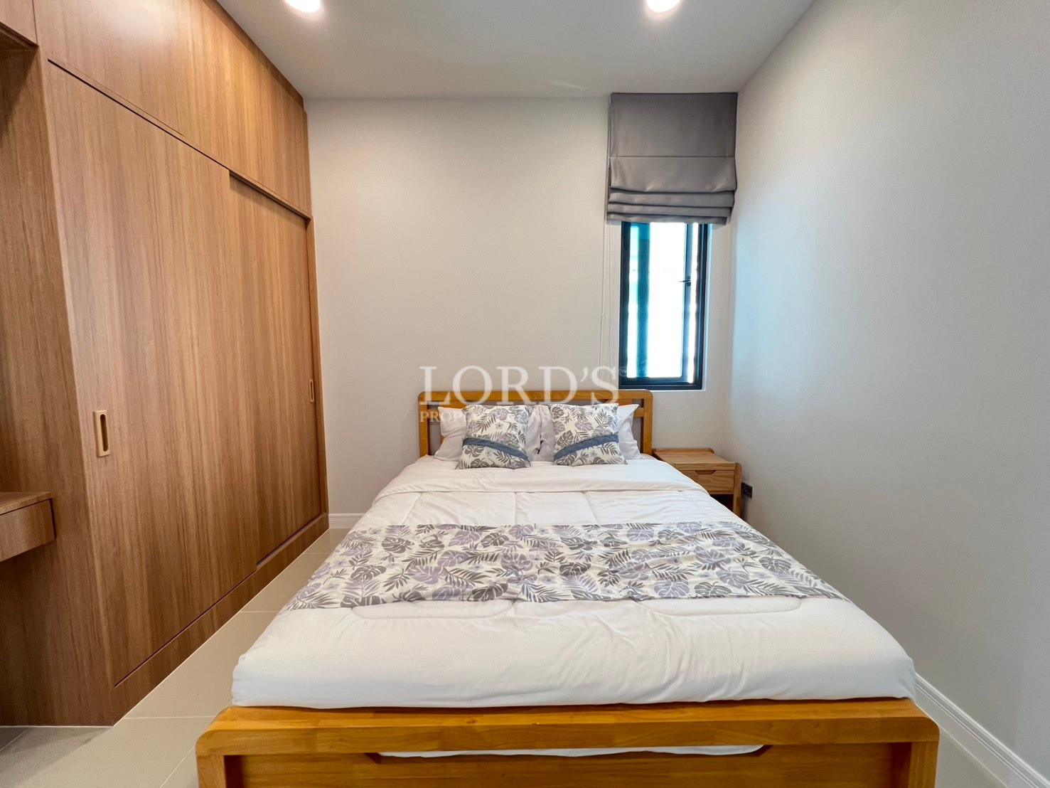 Compact modern bedroom with wooden bed, built-in wardrobe, and window with Roman blind.