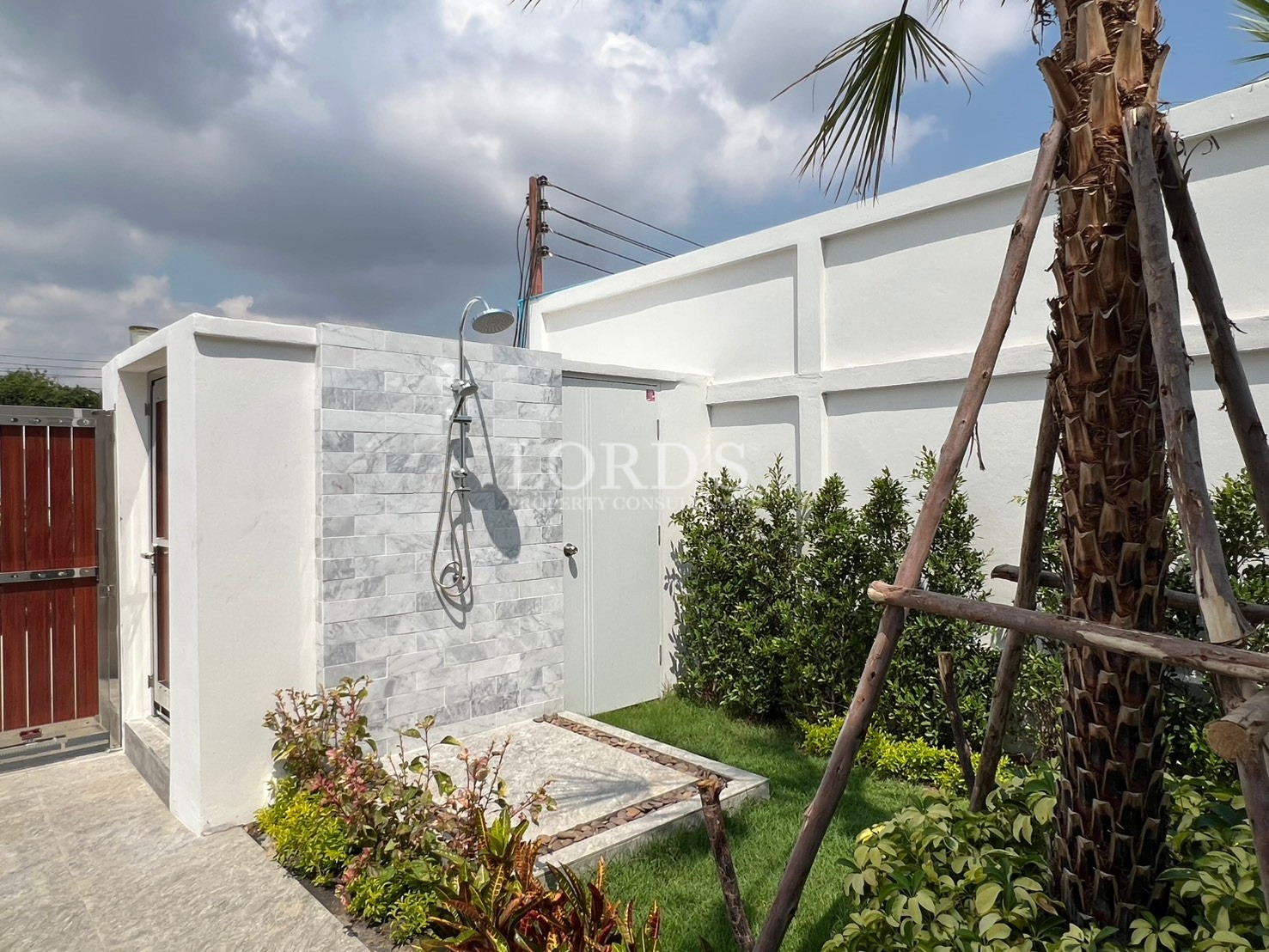 Outdoor garden shower with tiled feature wall, rain showerhead, and landscaped greenery beside a private boundary wall.