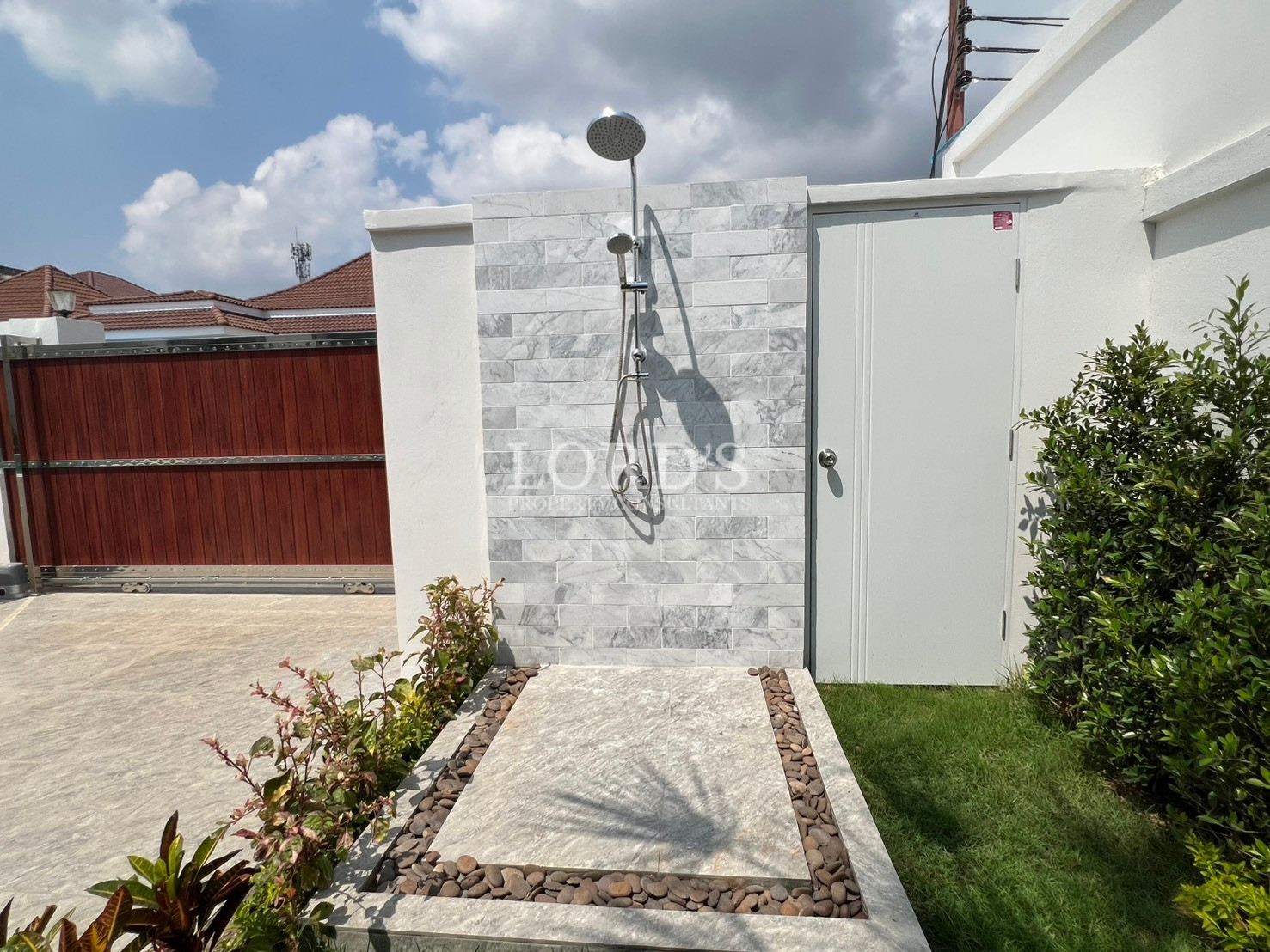 Modern outdoor shower with rain showerhead against a tiled wall, surrounded by landscaping and a private garden area.