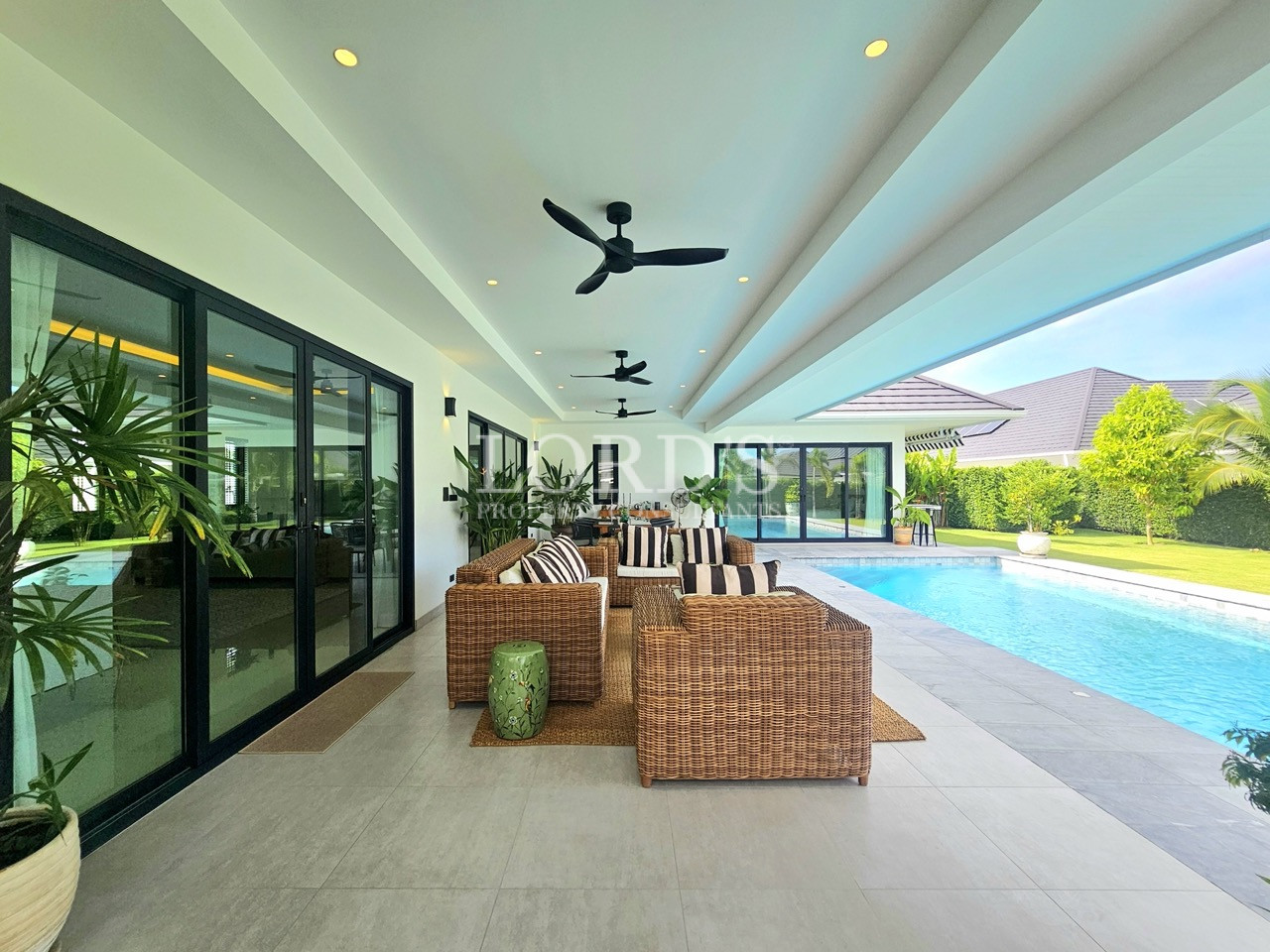 Covered poolside patio with wicker seating, ceiling fans, and tropical garden