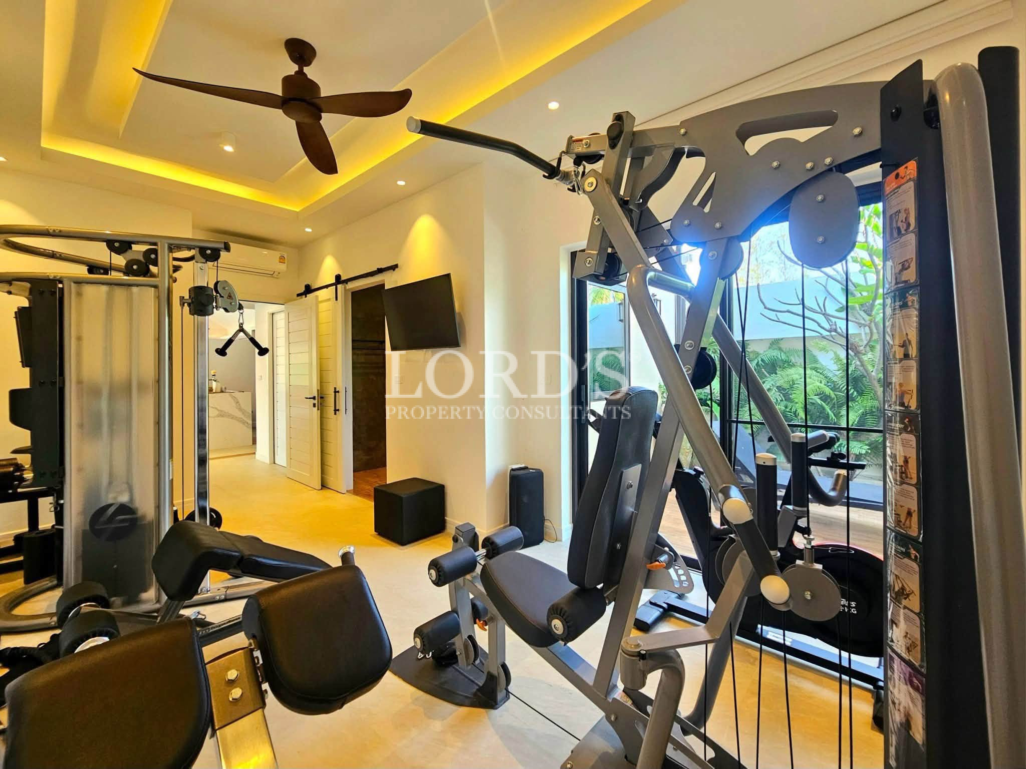 Modern private home gym with premium strength training equipment, cable machine, ceiling fan, and large windows overlooking a tropical garden