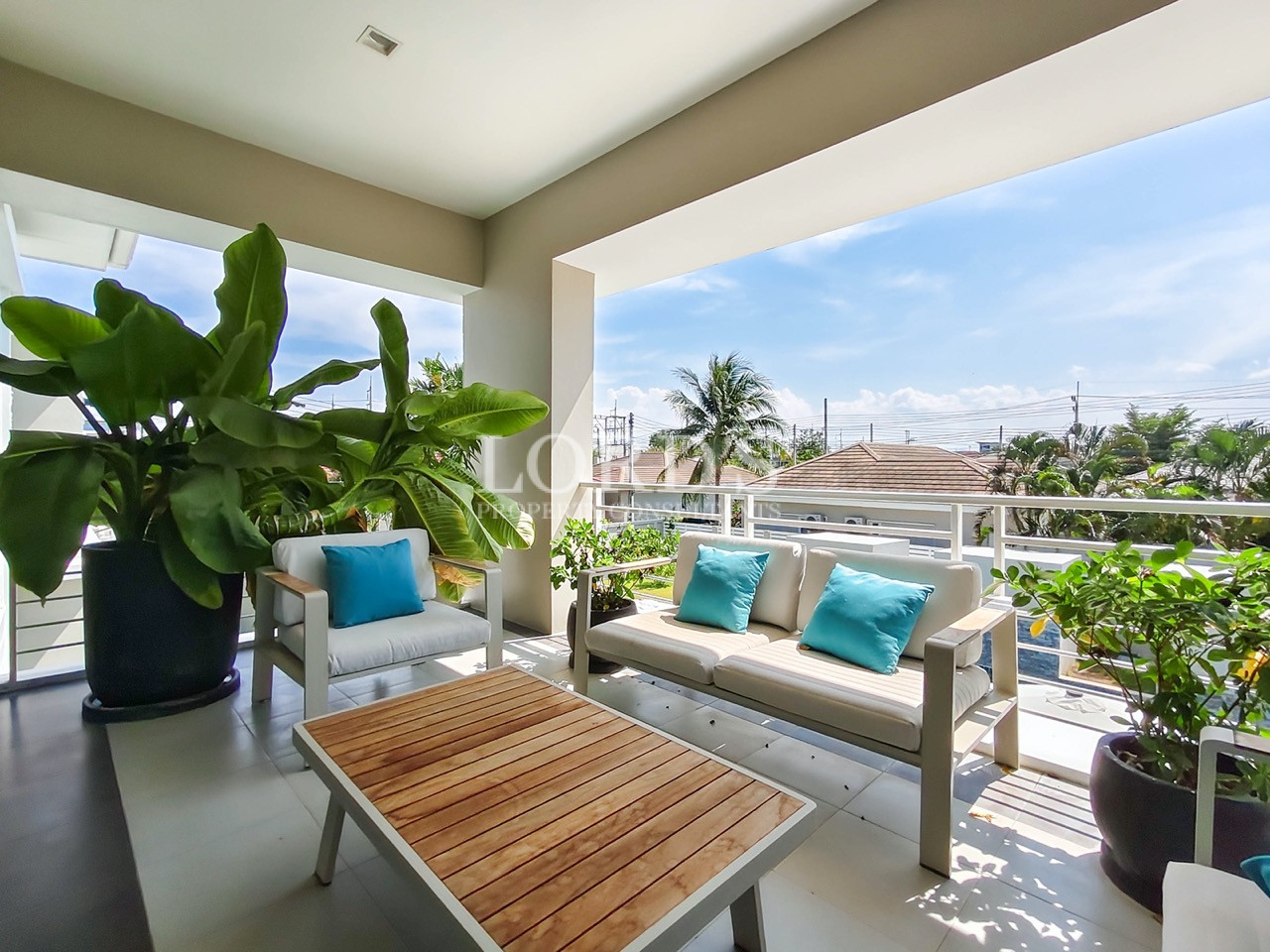 Luxury outdoor balcony with lounge seating, wooden coffee table, tropical plants, and panoramic neighborhood views