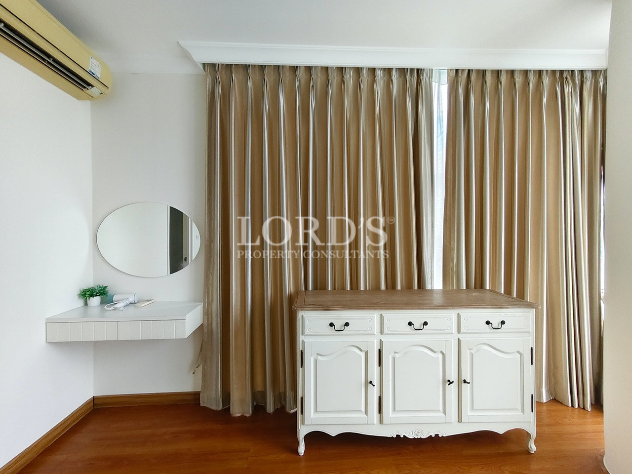 Bright bedroom corner with white dresser, oval mirror, beige curtains, and wooden flooring