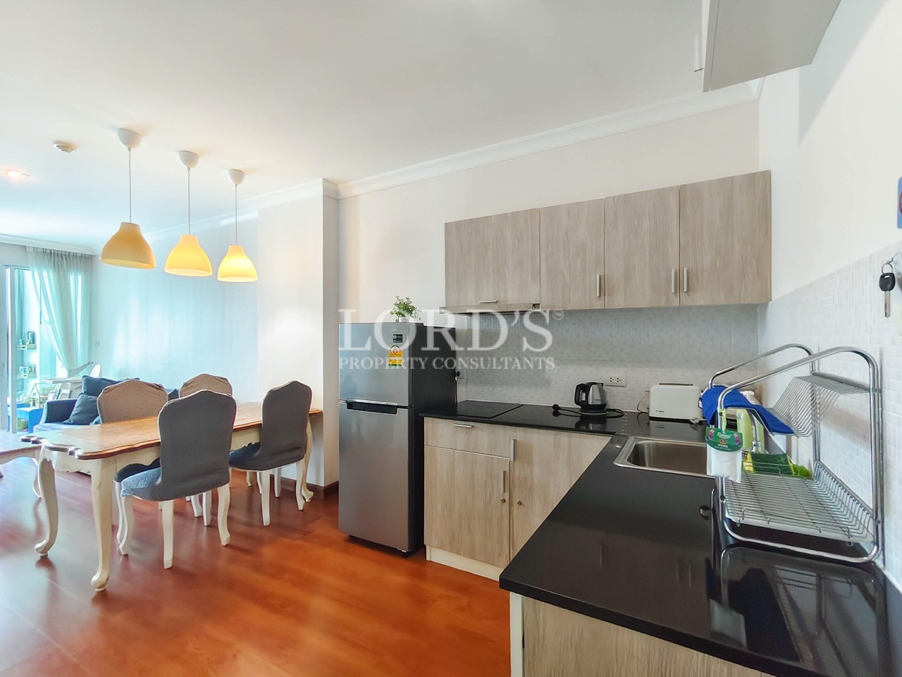 Modern apartment kitchen with light wood cabinets, black countertops, stainless steel refrigerator, and adjoining dining area