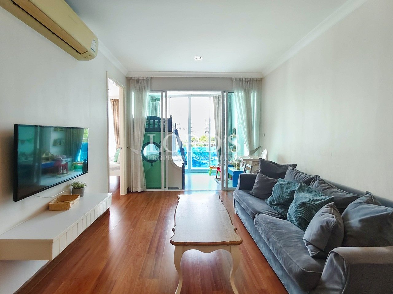 Modern living room with blue sofa, wooden flooring, wall-mounted TV, and pool-view balcony