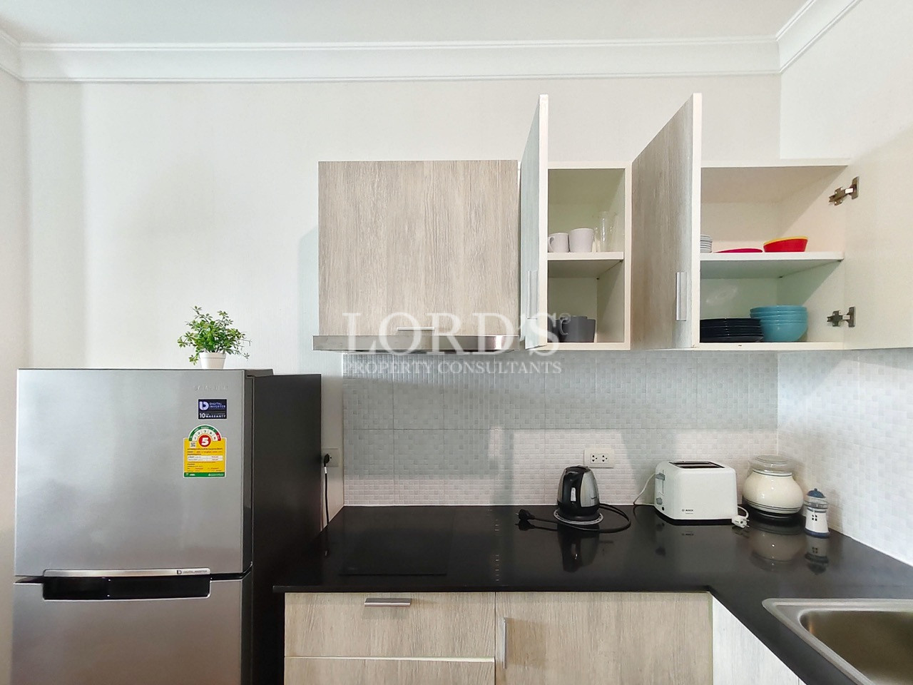 Modern apartment kitchen with light wood cabinets, black countertop, refrigerator, and open storage