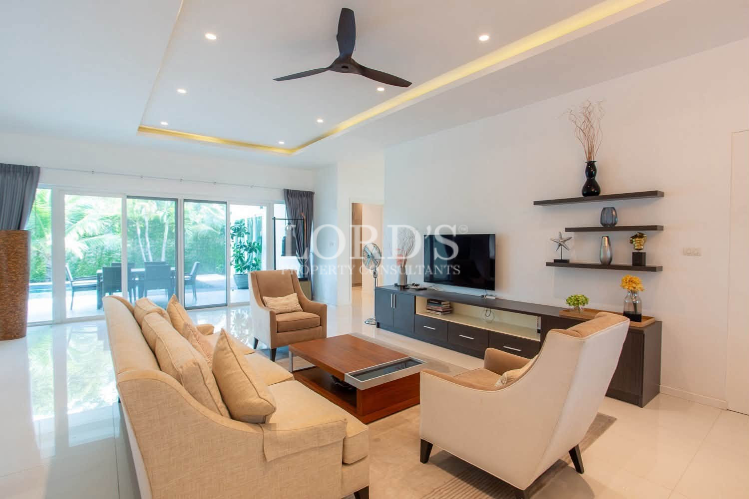 Bright living room with sofas, TV unit, ceiling fan, and large windows